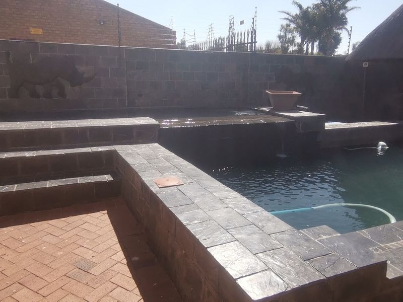 Outdoor pool area featuring a built-in water feature, stone-tiled pool surround, and brick paving, adjacent to a thatched roof structure.