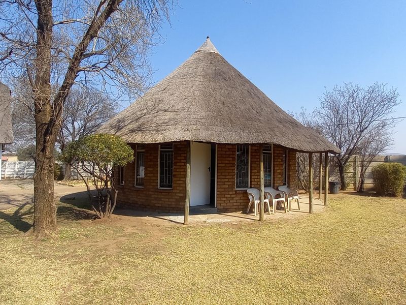 A detached circular dwelling featuring a traditional thatch roof, exposed brick exterior, and a covered veranda set within a spacious garden area.