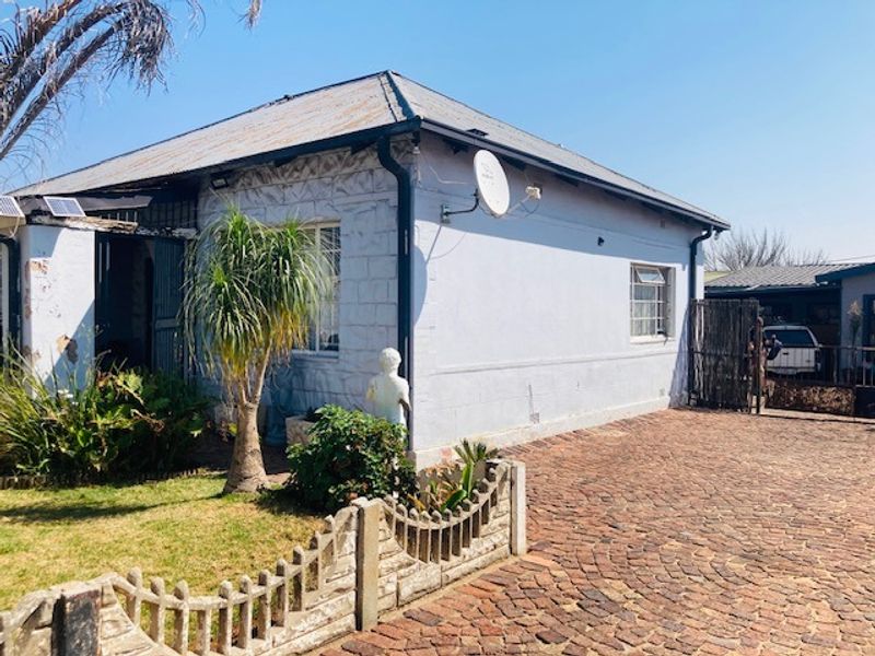 Front exterior view of a single-story house featuring a corrugated metal roof, brick paved driveway, and perimeter precast concrete fencing.