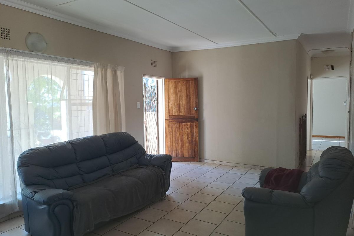 A living room featuring tiled flooring, a wooden entrance door with a security gate, and access to a hallway.