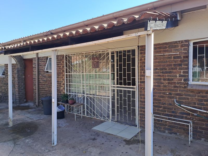 Rear exterior view featuring a covered patio area with brick facade, security gates, and concrete flooring, showing signs of wear and weathering.