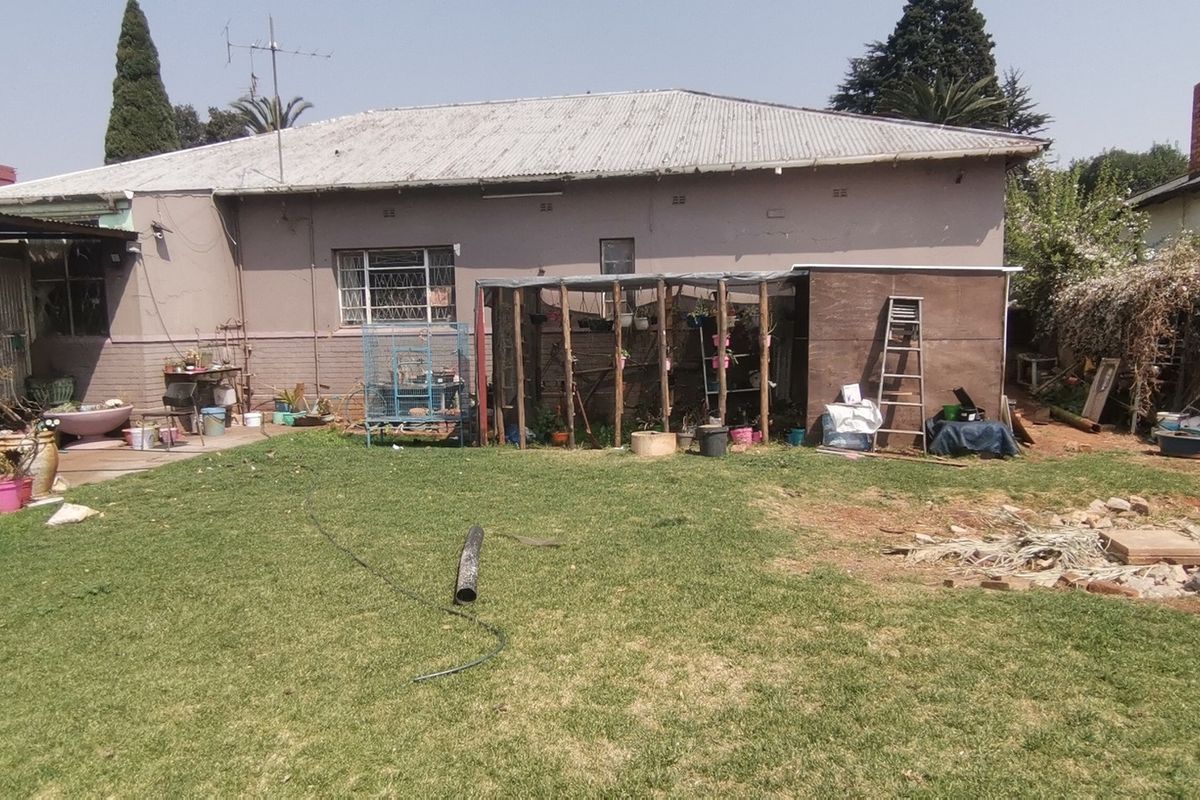 Rear view of the property featuring a corrugated metal roof, brick boundary wall, and a spacious garden area requiring maintenance.