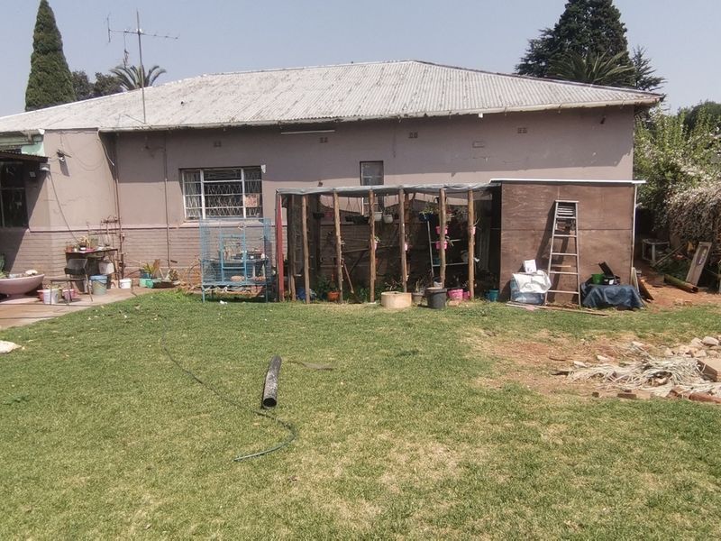 Rear view of the property featuring a corrugated metal roof, brick boundary wall, and a spacious garden area requiring maintenance.