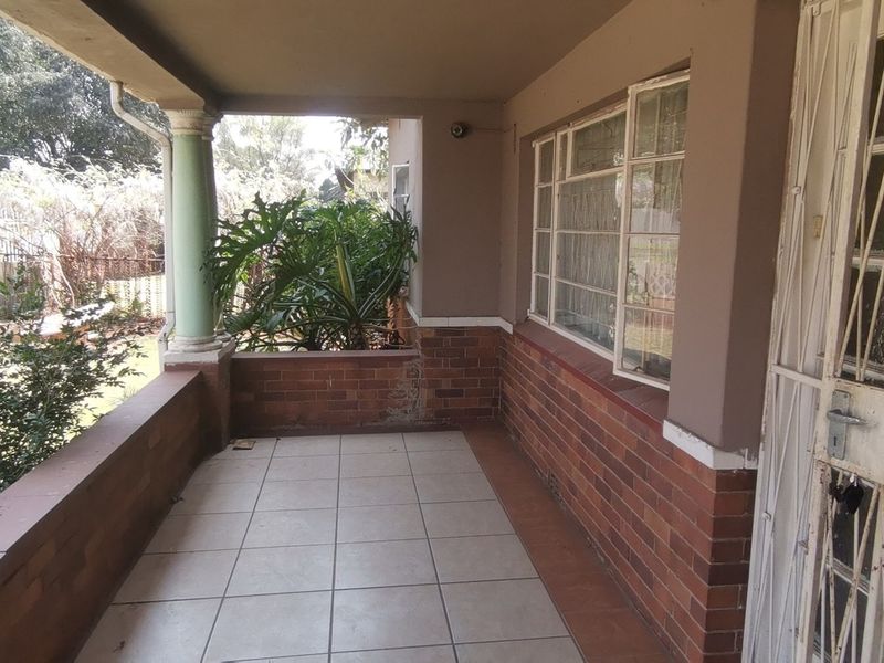 A covered front patio featuring tiled flooring, brick wall accents, and a metal security gate, overlooking the front garden area.