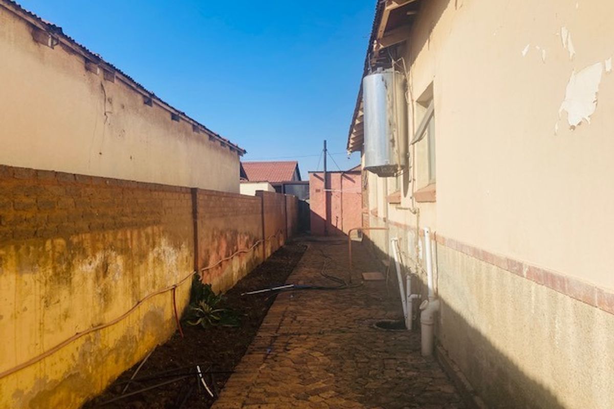 A narrow rear exterior walkway featuring a paved path, boundary wall, and external geyser unit with visible signs of weathering and paint deterioration.