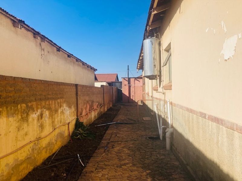 A narrow rear exterior walkway featuring a paved path, boundary wall, and external geyser unit with visible signs of weathering and paint deterioration.