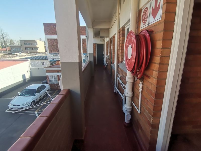 An exterior apartment building walkway featuring brick walls, a fire hose reel, and concrete flooring overlooking the parking area.