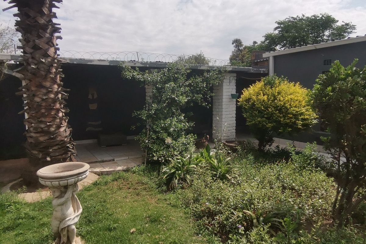 A rear garden area featuring a covered patio with brick pillars, mature landscaping, and perimeter security fencing.