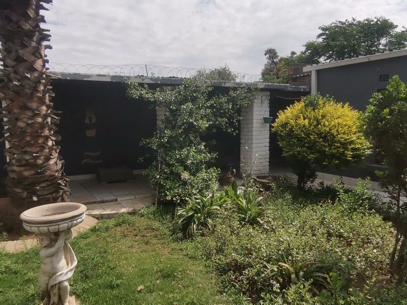 A rear garden area featuring a covered patio with brick pillars, mature landscaping, and perimeter security fencing.