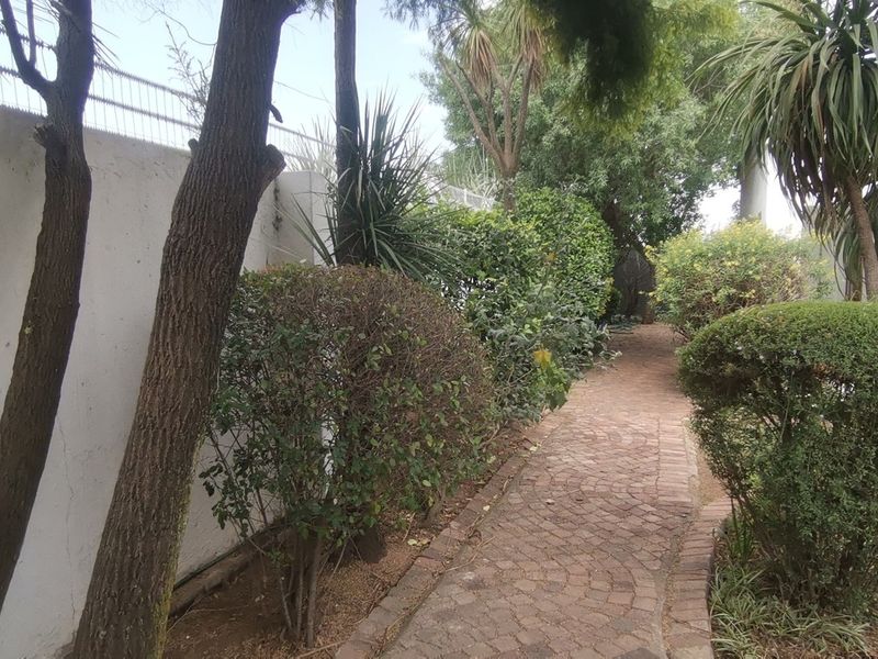 A landscaped garden area featuring a brick-paved pathway lined with mature trees and shrubs, enclosed by a perimeter boundary wall.