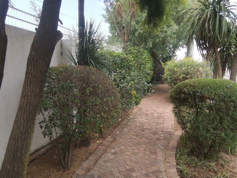 A landscaped garden featuring a brick paved pathway, mature trees, manicured shrubs, and a secure perimeter wall.