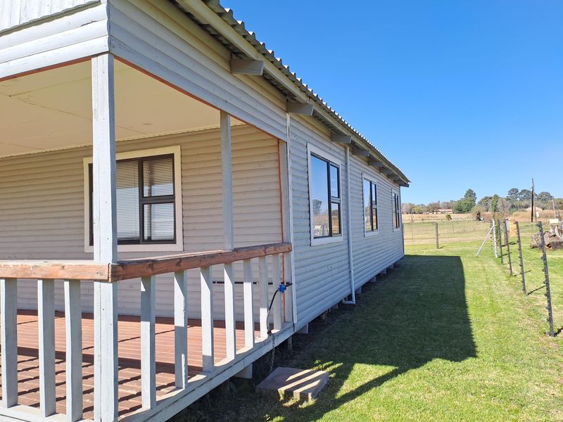 Side view of a timber-clad house featuring a covered wooden deck, aluminium windows, and a corrugated metal roof set on a rural property.