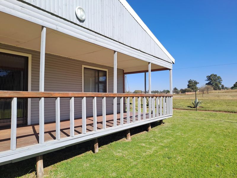 A covered timber deck patio featuring a wooden balustrade and sliding glass door access, overlooking a spacious rural garden area.