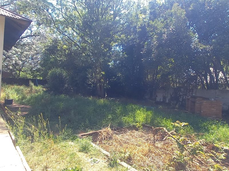 Rear exterior view of the property featuring a paved patio area, a built-in brick braai, and a large, overgrown garden space.
