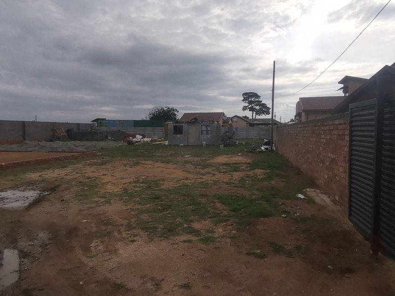 A large, undeveloped plot of land enclosed by perimeter brick walls and a metal gate, currently featuring unpaved ground and construction debris.