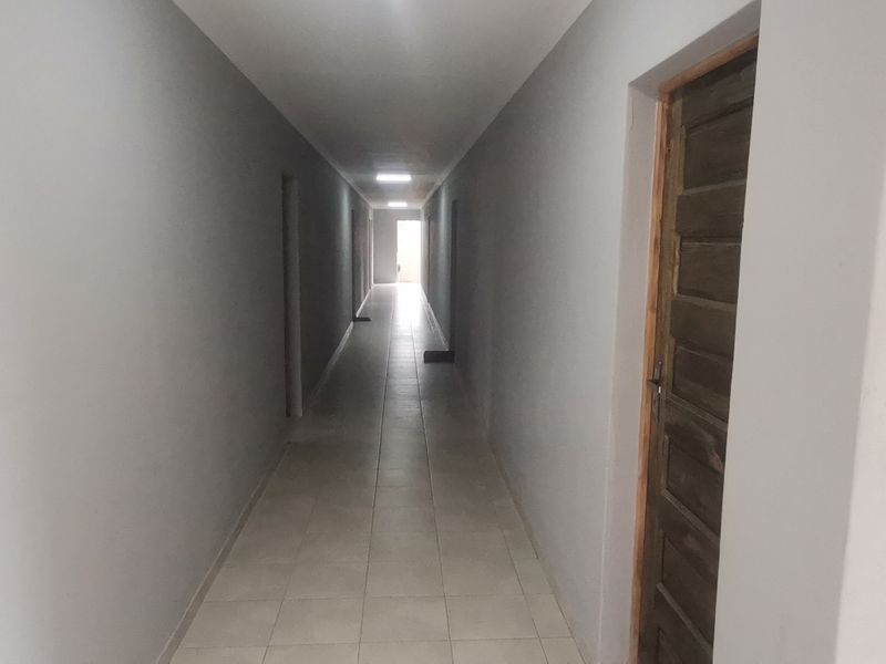 A long, empty hallway featuring tiled flooring, neutral grey walls, and recessed ceiling lighting, leading towards a bright doorway at the end.