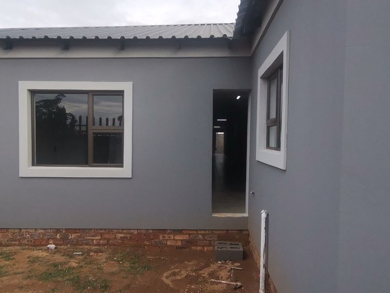 Rear exterior view of a modern grey plastered house featuring aluminium window frames and a corrugated metal roof, with unfinished surrounding ground.