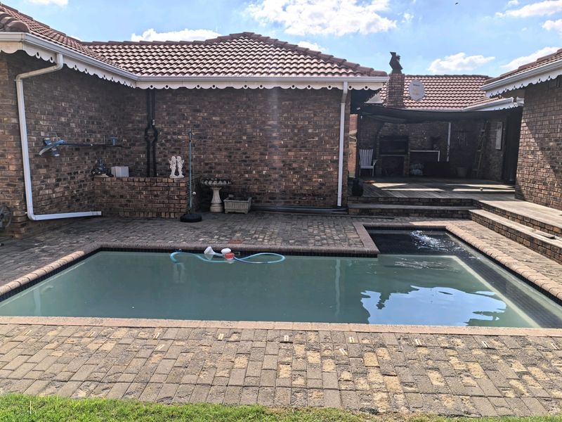 Outdoor pool area featuring brick paving, a built-in braai, and wooden decking, set against a face-brick house exterior.