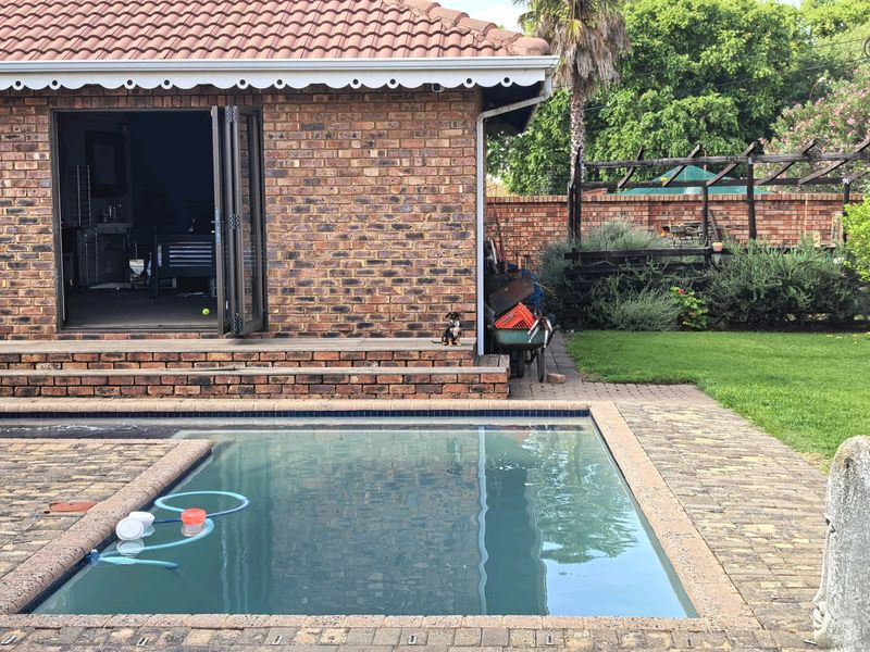 Outdoor pool area featuring a brick-clad dwelling with bi-fold doors, paved surround, and a rear garden with a timber pergola structure.