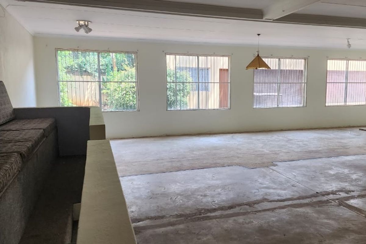 Spacious, open-plan living area featuring built-in concrete seating, large windows with security bars, and unfinished concrete flooring, offering significant potential for renovation.
