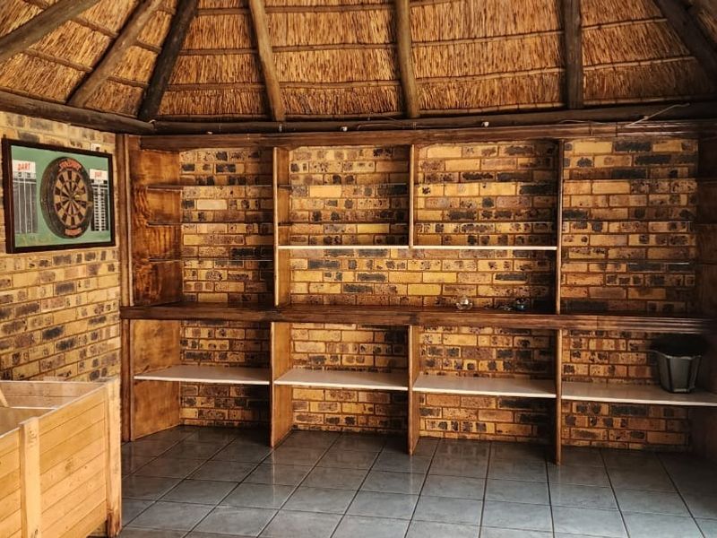 A rustic enclosed patio featuring a traditional thatched roof, exposed timber rafters, face brick walls, and extensive built-in timber shelving.