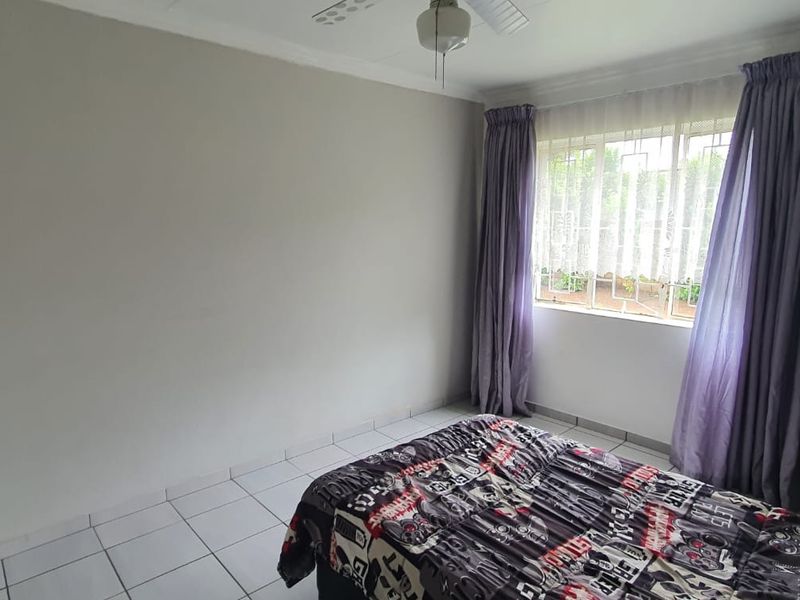 A bedroom featuring tiled flooring, a ceiling fan, and a built-in cupboard, with natural light entering through a window fitted with burglar bars.