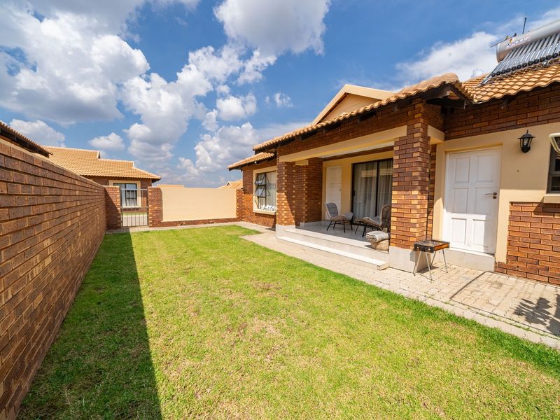 A well-maintained rear exterior view of a townhouse featuring a private lawn, brick boundary wall, paved patio area, and solar water heating system.