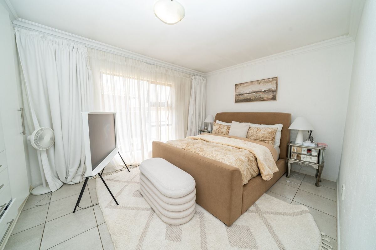A well-maintained bedroom featuring neutral tiled flooring, built-in cupboards, and large windows with floor-to-ceiling curtains allowing for ample natural light.