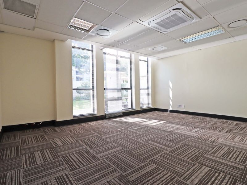 A bright, empty office space featuring carpet tiles, a suspended ceiling with integrated lighting, air conditioning, and large windows providing natural light.