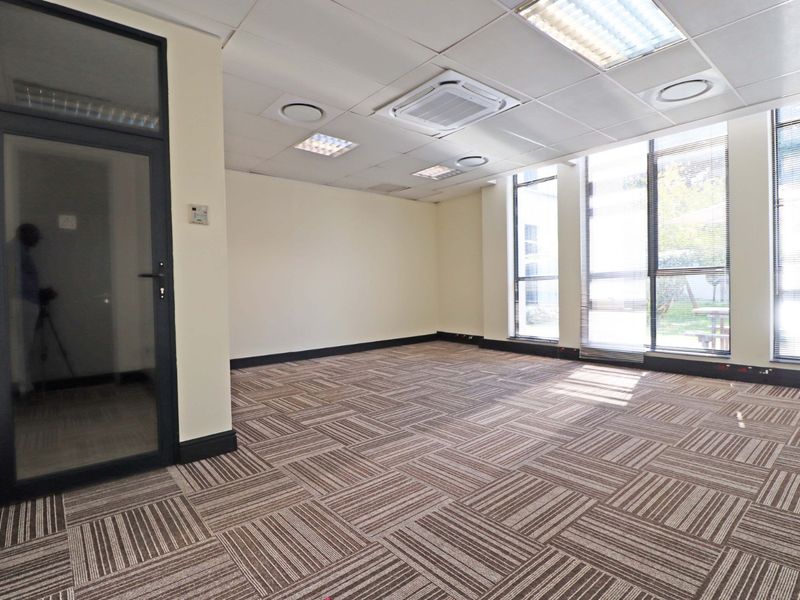 A bright, vacant office space featuring carpet tiles, a suspended ceiling with recessed lighting, integrated air conditioning, and large windows providing natural light.