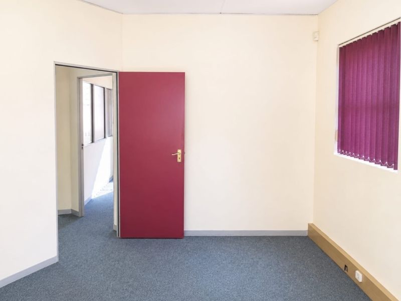 A clean, empty office space featuring neutral walls, grey carpet flooring, and vertical window blinds, providing a functional environment for professional use.