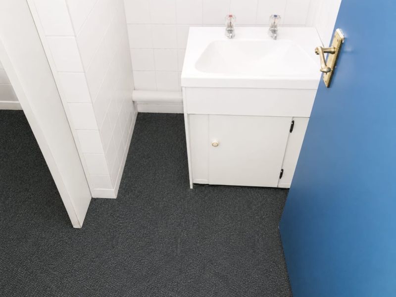 A functional office bathroom featuring a white vanity basin with tiled splashback and dark grey carpeting.
