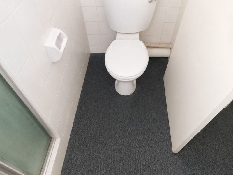 A compact office bathroom featuring a standard toilet, white tiled walls, and dark carpeting.