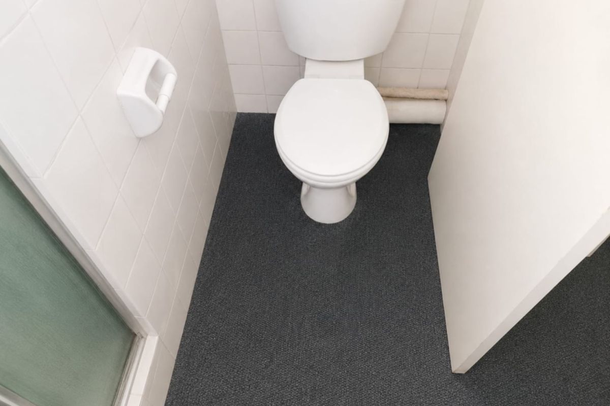 A compact office bathroom featuring a standard toilet, white tiled walls, and dark carpeting.