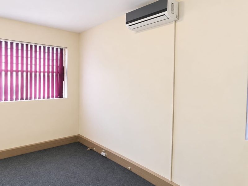 A clean, empty office space featuring neutral walls, grey carpet, vertical blinds, and a wall-mounted split-system air conditioning unit.
