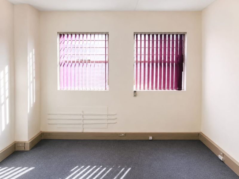 A vacant office space featuring neutral walls, grey carpeting, and dual windows fitted with vertical blinds.