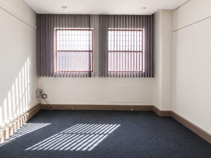 A bright, empty office space featuring dark carpeting, neutral walls, and vertical blinds on dual windows, providing a clean and functional workspace.