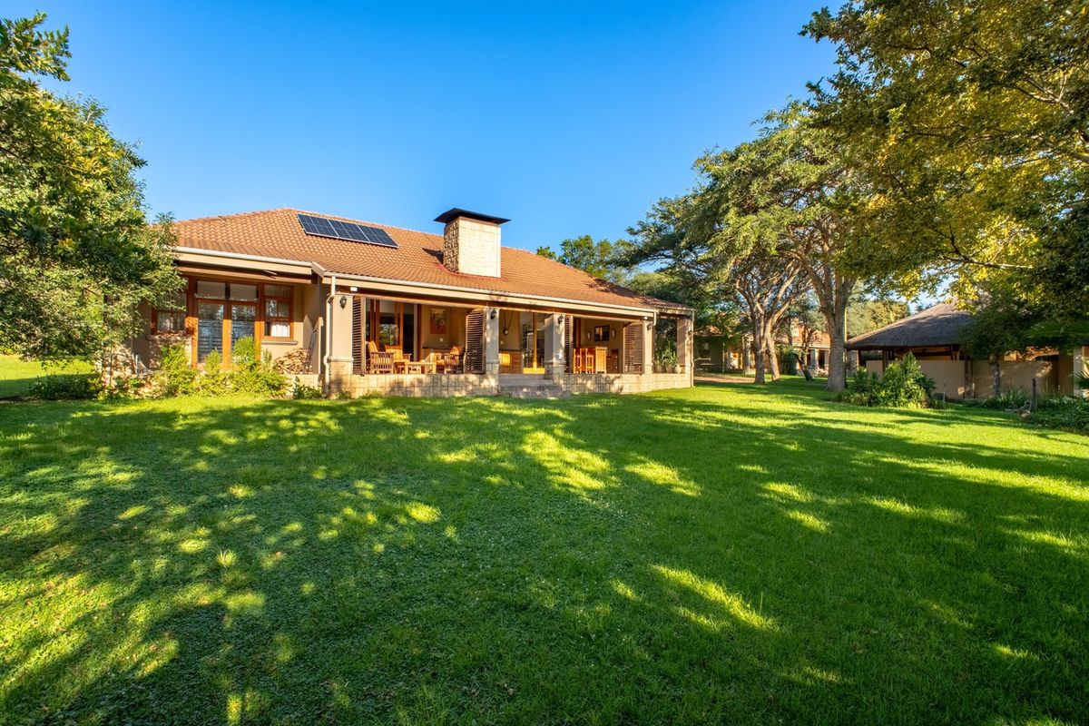 Rear view of a well-maintained farm-style residence featuring a spacious covered patio, stone chimney, and solar panels set within a lush green garden.