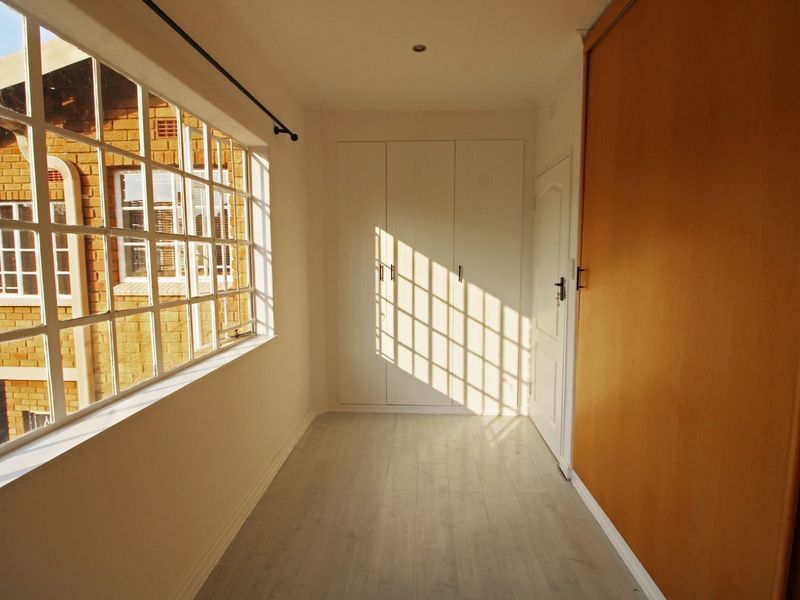 A bright, empty bedroom featuring light laminate flooring, built-in cupboards, and a large window allowing for ample natural light.