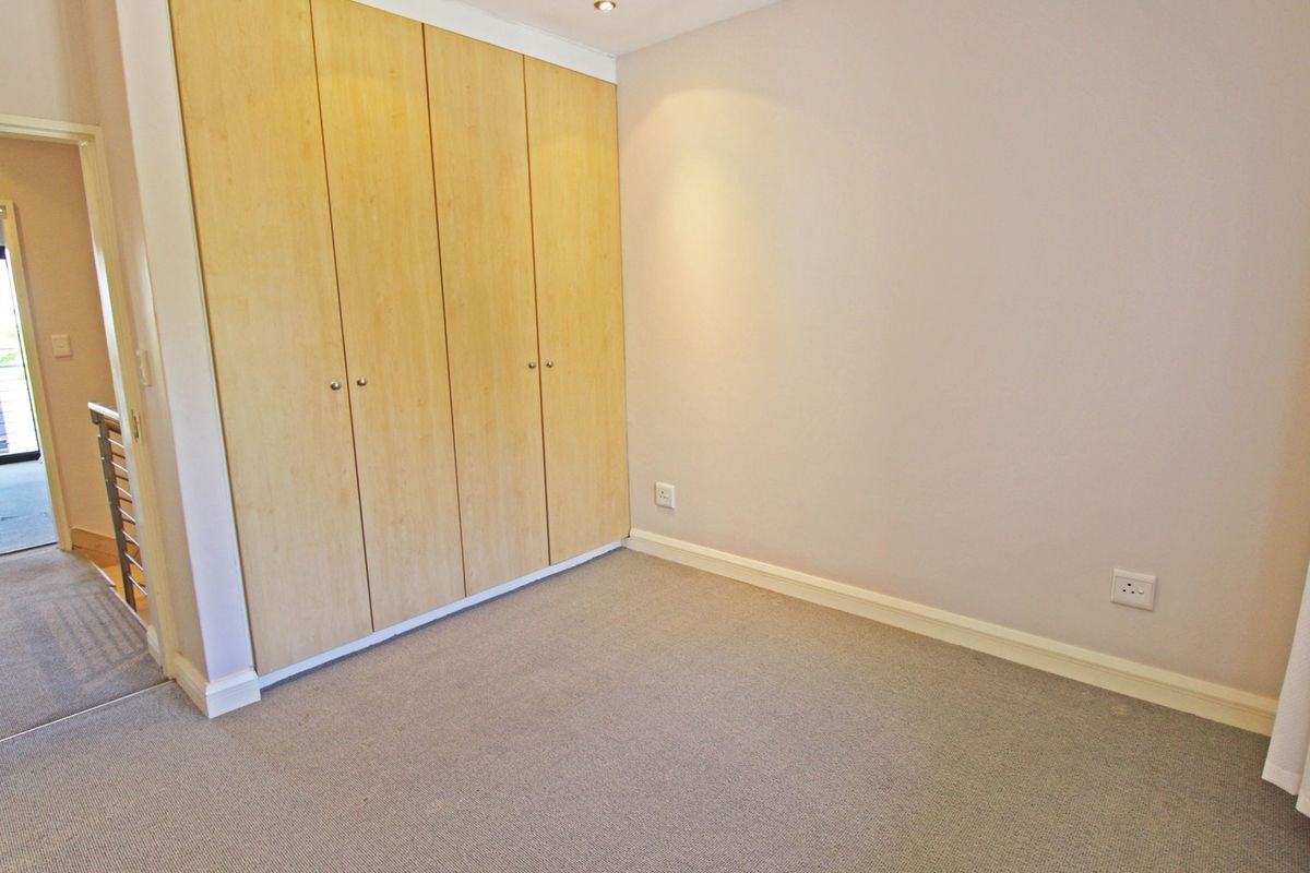 A clean, empty bedroom featuring built-in wooden cupboards, neutral carpeting, and recessed ceiling lighting, with an adjacent hallway visible.