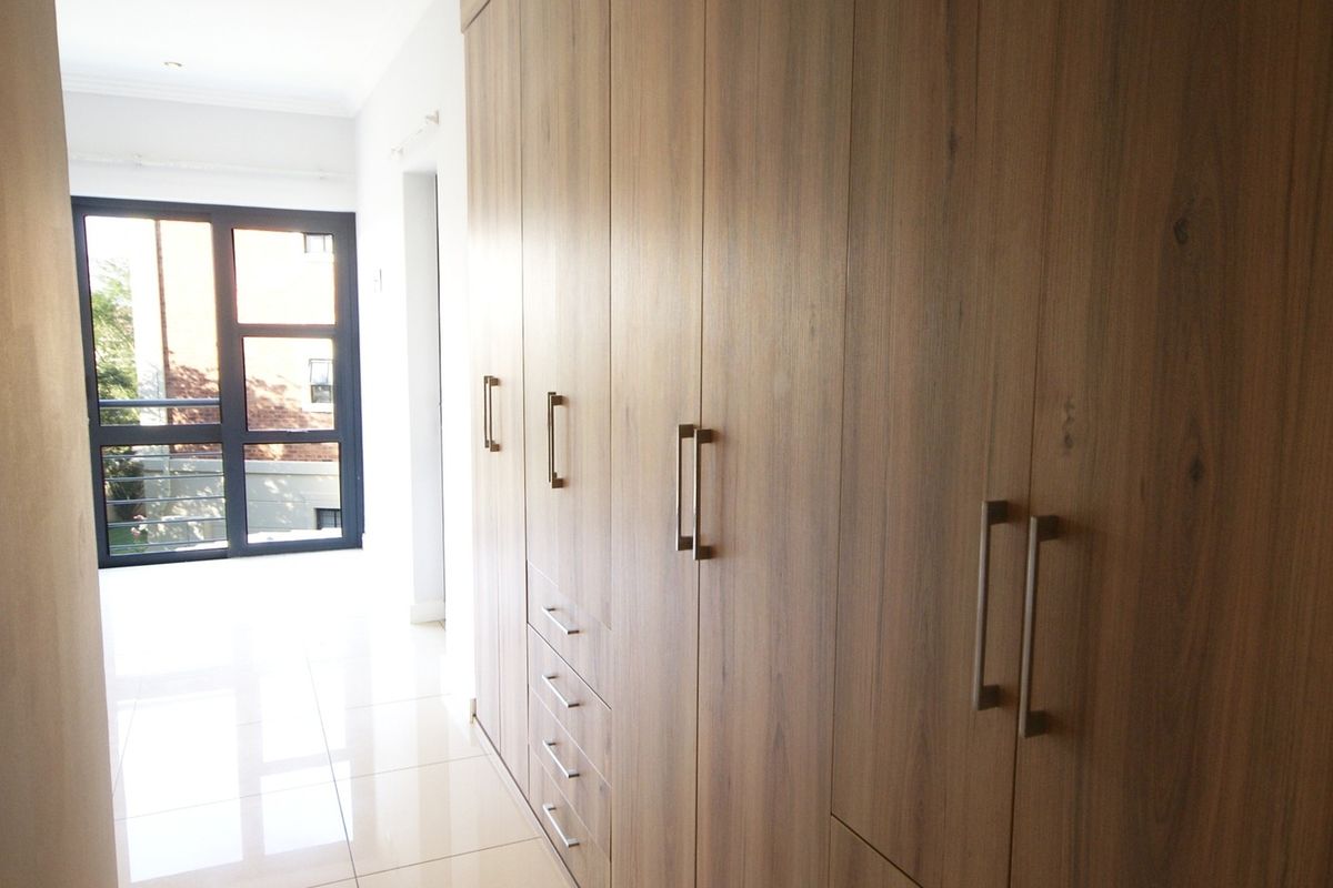 A bright bedroom featuring extensive floor-to-ceiling built-in wooden cupboards, polished tile flooring, and a large window providing natural light.