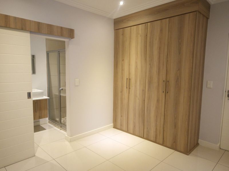 A clean, modern bedroom featuring light wood-finish built-in cupboards, tiled flooring, and direct access to an en-suite bathroom.