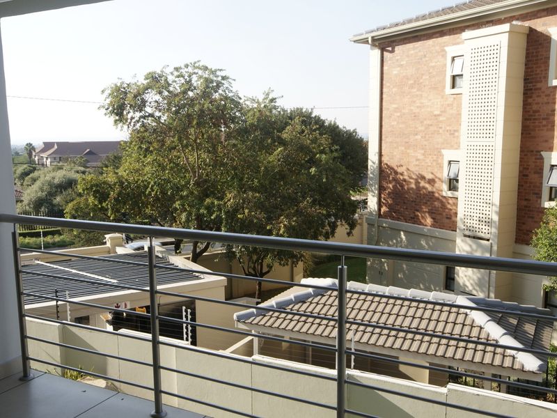 A covered balcony featuring a modern stainless steel balustrade and tiled flooring, overlooking the surrounding residential complex and garden area.