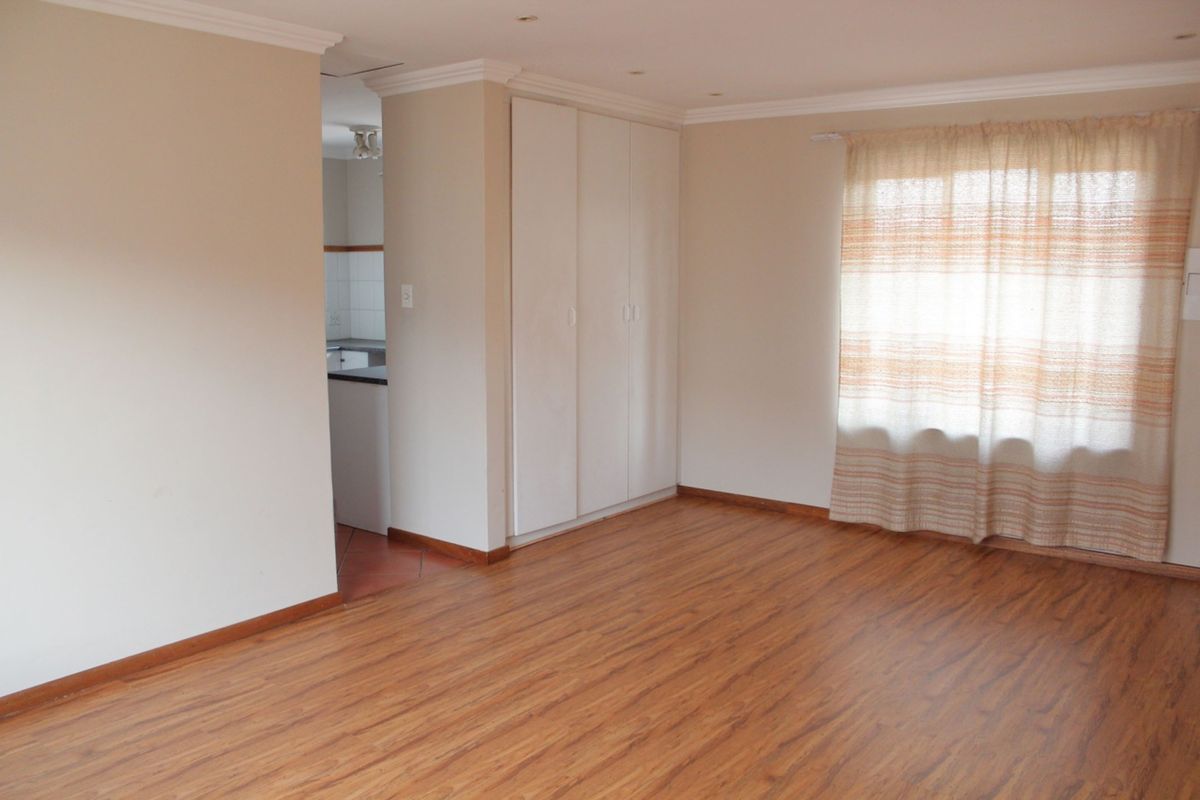 A bright, empty bedroom featuring laminate flooring, built-in cupboards, and neutral wall finishes, with a view into the adjacent kitchen area.