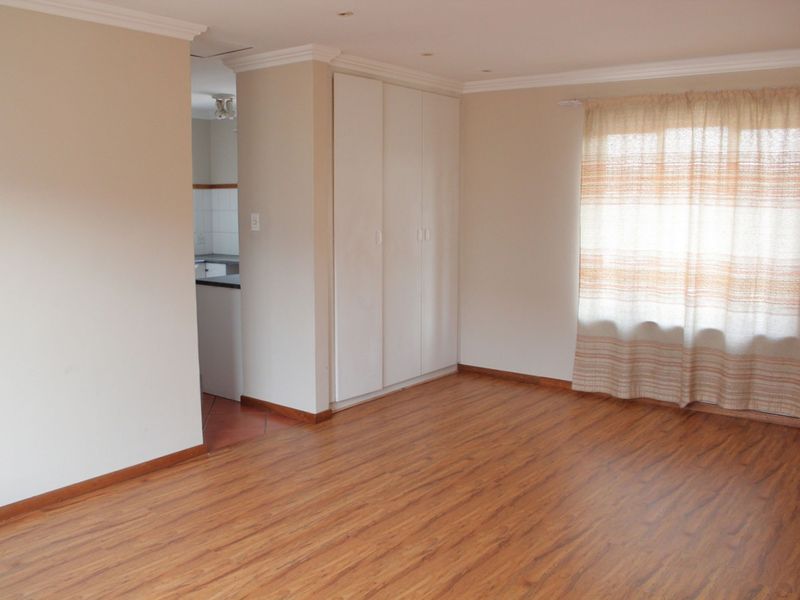 A bright, empty bedroom featuring laminate flooring, built-in cupboards, and neutral wall finishes, with a view into the adjacent kitchen area.