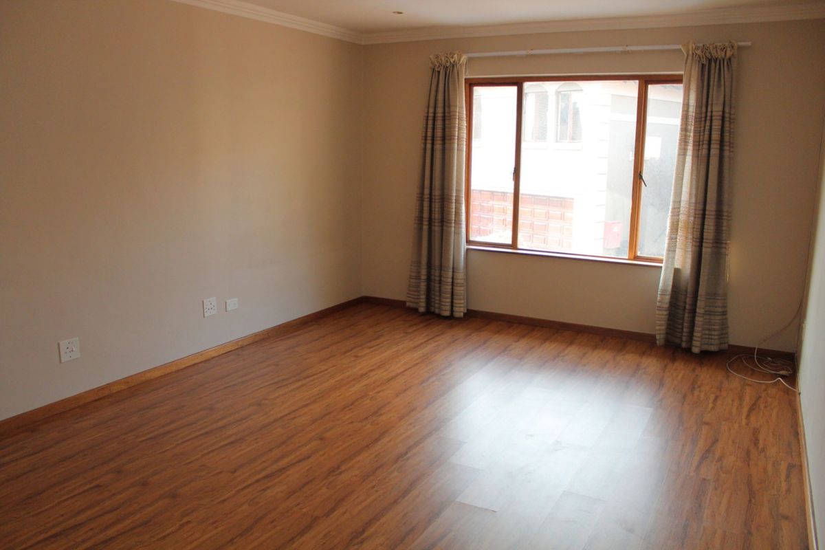 A bright, empty living room featuring warm-toned laminate flooring, neutral walls, and a large window with timber frames.