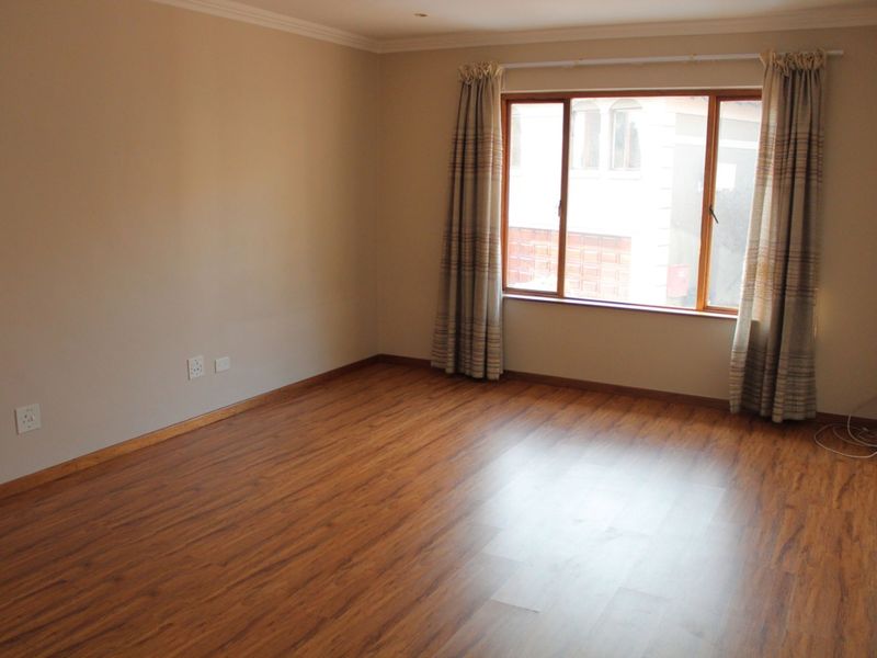 A bright, empty living room featuring warm-toned laminate flooring, neutral walls, and a large window with timber frames.