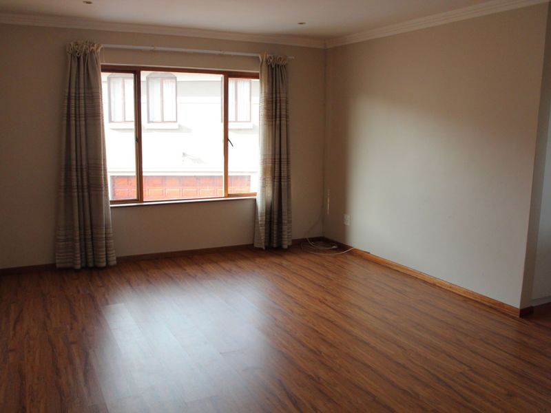 A spacious, empty living room featuring warm-toned laminate flooring, recessed downlighting, and a large window with neutral curtains.