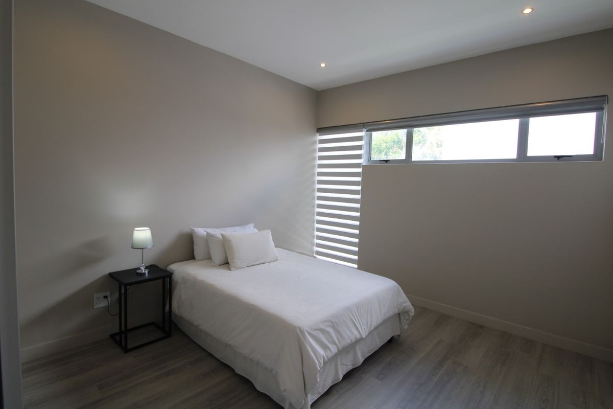 A modern bedroom featuring neutral wall tones, laminate flooring, recessed downlights, and a horizontal window fitted with zebra blinds.