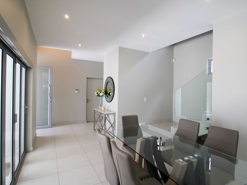 Modern open-plan living area featuring neutral tiled flooring, recessed lighting, and a contemporary glass balustrade staircase, leading to outdoor access via aluminium sliding doors.